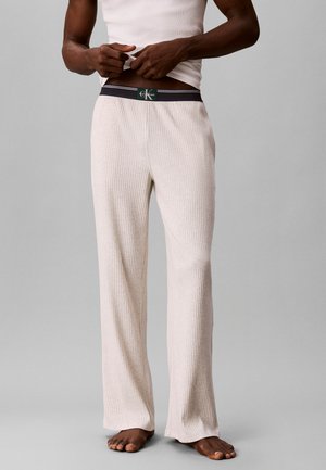 Uomo che indossa pantaloni in maglia crema a coste con fascia nera Calvin Klein, abbinati a una maglietta bianca senza maniche, in piedi a piedi nudi su pavimento grigio.