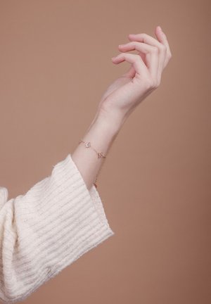 Růžově zlatý náramek s malým srdcem a diamantovými akcenty, nošený na ruce s texturovaným krémovým svetrem, na béžovém pozadí.