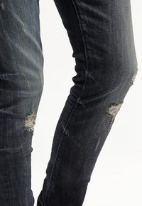 Jean en denim noir usé avec des patchs usés aux genoux et aux cuisses sur fond blanc.