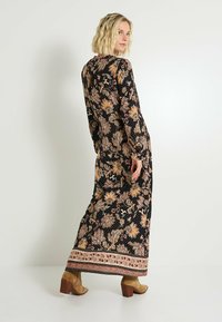 Robe noire longue avec un motif floral complexe en tons dorés et roses. Présente des manches longues et un ourlet à motif. Portée avec des bottes à talons couleur fauve.