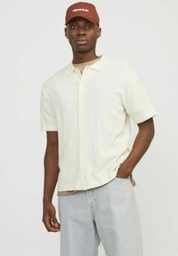 Kreemivärviline polo-särk, millel on nupukinnitus, lühikesed varrukad ja lõdvestunud istuvus, koos helehallide teksade ja pruuni mütsiga.