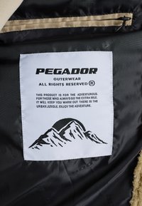 Balta auduma etiķete ar melnu tekstu un kalnu grafiku. Teksts ietver "PEGADOR OUTERWEAR" un produkta aprakstu melnā fontā.