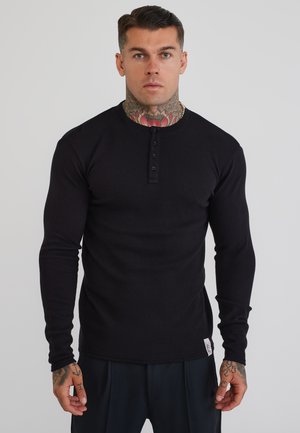 Uomo con tatuaggi al collo e sulla mano che indossa una maglietta henley nera aderente a manica lunga, in piedi davanti a uno sfondo chiaro e uniforme.