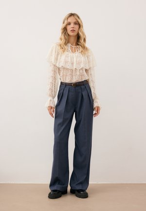 Biała koronkowa bluzka z falbankami na rękawach i detalem w postaci wiązania, zestawiona z ciemnoniebieskimi, wysokimi spodniami o szerokich nogawkach oraz czarnymi butami.