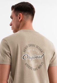 Béžové bavlněné tričko s kulatým výstřihem a krátkými rukávy. Na zadní straně se nachází potištěné logo se slovem "Original" a dekorativními textovými prvky.