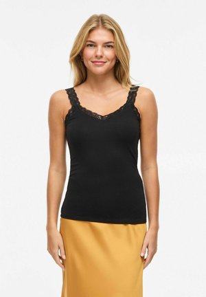 Femme blonde portant un caraco noir orné de dentelle et une jupe jaune moutarde, debout devant un fond blanc uni, souriant doucement.