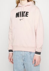 Sudadera rosa claro hecha de una tela suave, con un gran logo negro de "NIKE" en la parte frontal y un bolsillo delantero. Puños con detalles en gris.