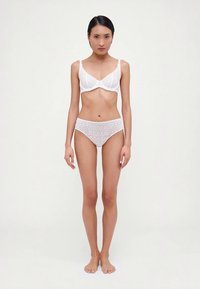 Witte kanten beha met geschulpte randen en bijpassende kanten bikini. Beide hebben bloemmotieven en een zachte textuur. Model staat op blote voeten.