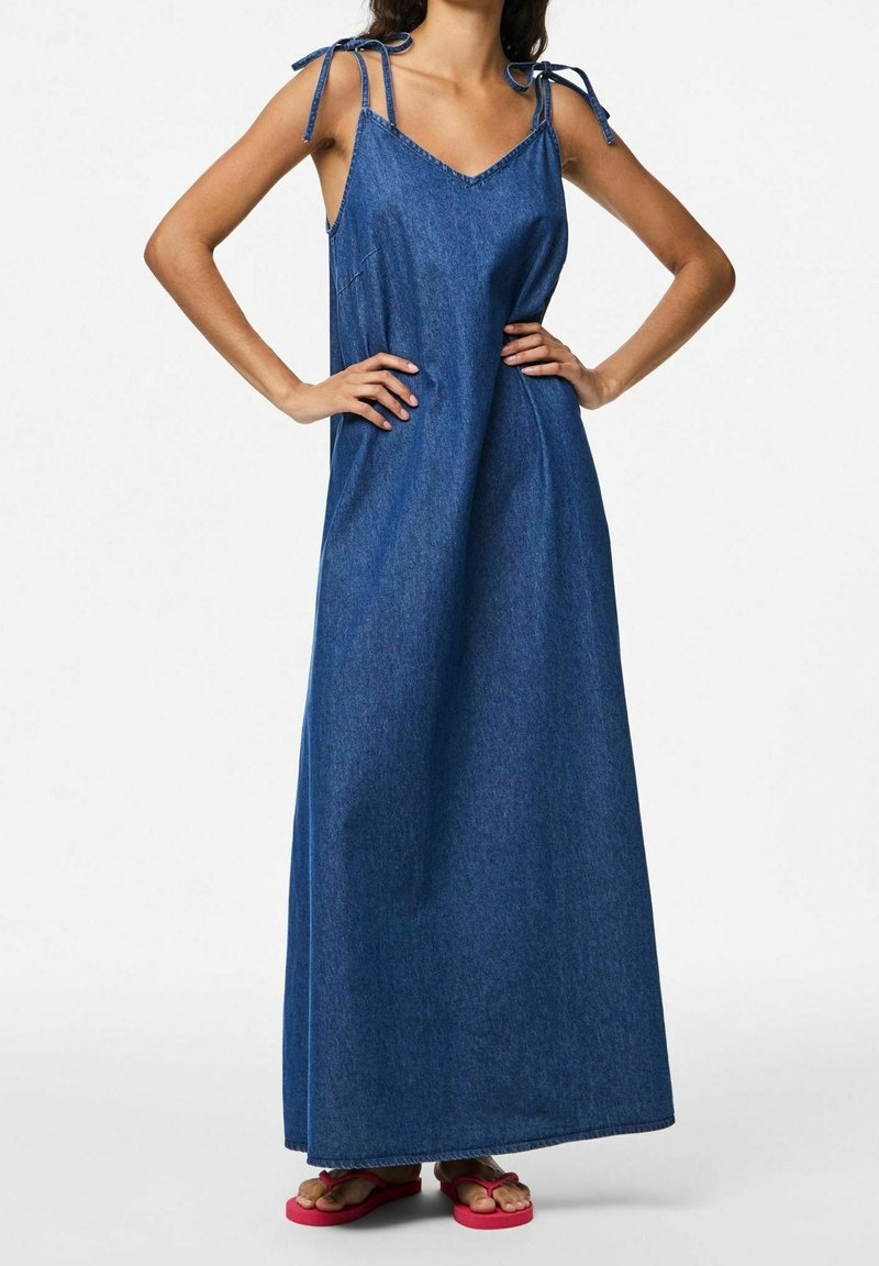Femme portant une robe maxi en jean bleu sans manches avec des bretelles nouées aux épaules et des tongs rouges, debout les mains sur les hanches.