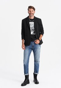 Svart kavaj över en grafisk t-shirt, slitna ljusblå jeans och svarta snörboots. Har en avslappnad look med lager av accessoarer.