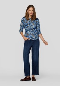 Blaue, floral gemusterte Bluse mit langen Ärmeln und rundem Ausschnitt, kombiniert mit weit geschnittenen Jeans aus dunklem Denim und braunen Slippern.