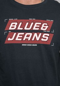 T-shirt en coton noir avec des graphismes audacieux en rouge et blanc portant l'inscription "BLUE & JEANS", avec des accents rayés et des détails de marque imprimés.