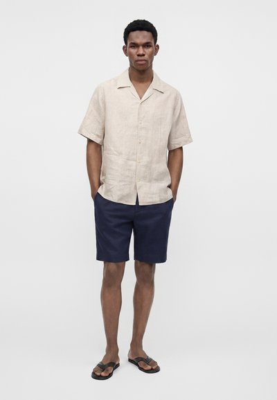 Homme debout, les mains dans les poches, portant une chemise beige à manches courtes boutonnée, un short bleu marine et des sandales noires sur fond blanc.