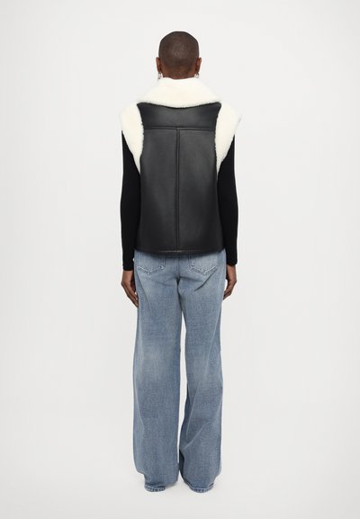 Black leather vest with white shearling collar, sleeveless design, and fitted shape. Worn over a long-sleeve black top and blue wide-leg jeans.
