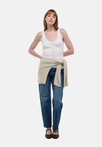 Débardeur blanc avec motif en maille verticale, pull beige noué à la taille, jean bleu à coupe ample et chaussures tissées marron. Fond uni.