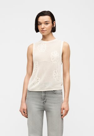 Γυναίκα με κοντά σκούρα μαλλιά φοράει λευκό αμάνικο τοπ από δίχτυ με floral μοτίβα και ανοιχτό γκρι τζιν, στέκεται μπροστά σε απλό φόντο.