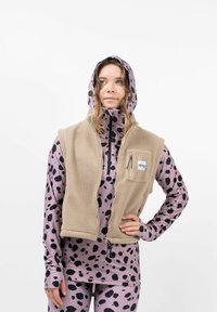 Beige Fleece-Weste mit Reißverschluss-Taschen, getragen über einem rosafarbenen Oberteil mit schwarzem gepunktetem Muster. Die Kapuzenform erhöht den Stil und die Wärme.