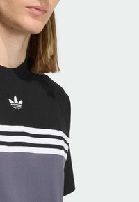 Čierno-sivé tričko s krátkym rukávom s bielymi vodorovnými pruhmi a malým logom na hornej časti hrudníka. Mäkká textúra látky.