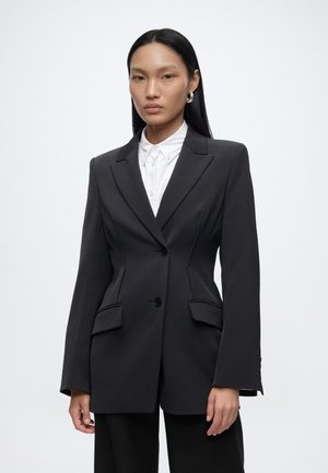 Mujer con largo cabello negro lleva una camisa blanca abotonada y un blazer negro a medida sobre un fondo blanco liso.