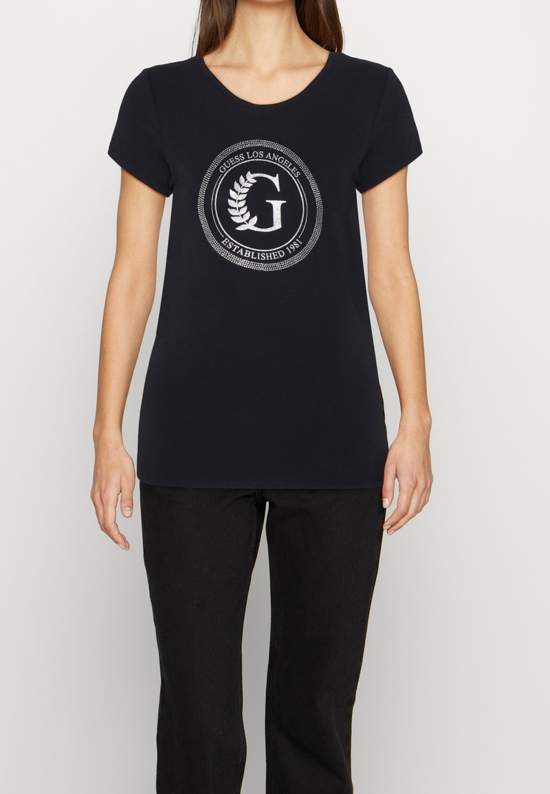 Sort t-shirt med korte ærmer og rund halsudskæring, der har trykt et sølvlogo med en laurbærkrans og teksten "Guess Los Angeles Established 1981."