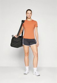 Pomarańczowa obcisła koszula z krótkim rękawem, czarne sportowe spodenki z białym obszyciem, noszący czarną torbę podróżną. Białe sneakersy i skarpetki.