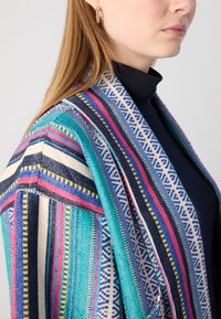 Cardigan tissé multicolore avec des rayures turquoise, marine et rose, des motifs complexes et une coupe décontractée, porté sur un col roulé noir.