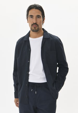 Camisa de botones azul marino con cuello, un bolsillo en el pecho y puños enrollados, sobre una camiseta blanca y combinada con pantalones oscuros.