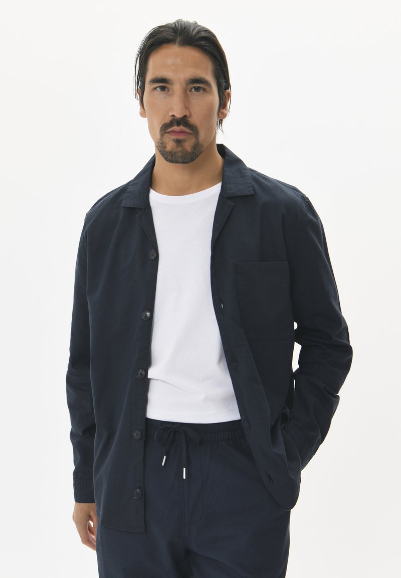 Camisa de botones azul marino con cuello, un bolsillo en el pecho y puños enrollados, sobre una camiseta blanca y combinada con pantalones oscuros.