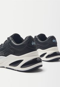 Sneakers nere in pelle con una spessa suola bianca e un accento nero lucido. La fodera distintiva e il marchio sul tallone aggiungono dettagli.