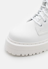Botín de cuero blanco hasta el tobillo con una suela de plataforma gruesa y texturada y cordones blancos. Presenta ojales redondos y sutiles detalles de costura.