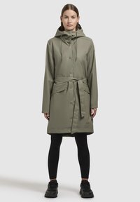 Olive green waterproof trench coat with a belted waist, hood, snap buttons, and two front pockets. Worn over black leggings and sneakers.