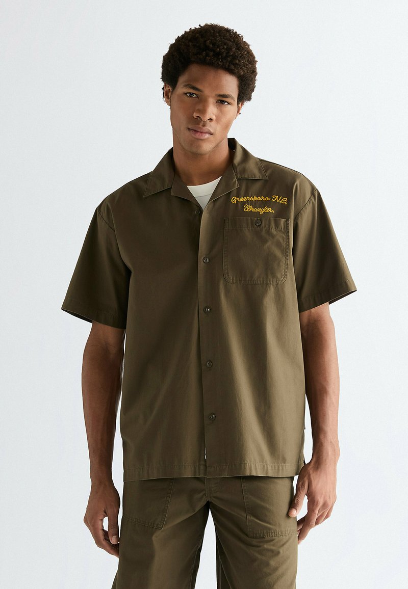 Olivengroen shirt met korte mouwen, een knoopsluiting aan de voorkant, een zakje en gele geborduurde tekst met de woorden "Greensboro NC Wrangler." Gladde textuur.