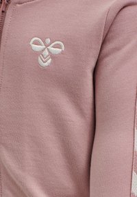 Różowa bluza z teksturowanej tkaniny; na piersi haftowane logo pszczoły w białym kolorze oraz kontrastowy akcent w wzór na rękawie.