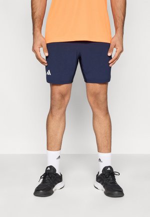 Marineblaue Sportshorts mit strukturierter Oberfläche und weißem Adidas-Logo. Kombiniert mit einem leuchtend orangefarbenen Shirt und schwarzen Sneakers.