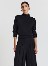 Pull noir à col montant avec des manches longues, fabriqué en tissu doux. Associé à un pantalon large noir à plis. Design simple et monochrome.