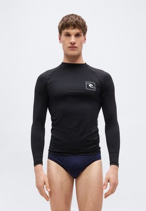 Νεαρός άνδρας που φοράει μαύρο μακρυμάνικο αντηλιακό μπλουζάκι με λογότυπο και μπλε σκούρα μαγιό, στέκεται μπροστά σε μια απλή ανοιχτόχρωμη φόντο.