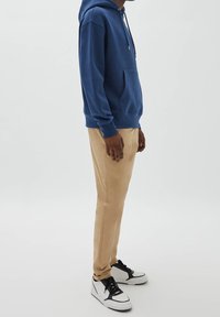 Blauer Hoodie-Pullover mit Vordertasche, kombiniert mit beigen, zulaufenden Hosen und weißen Sneakern mit schwarzen Akzenten.