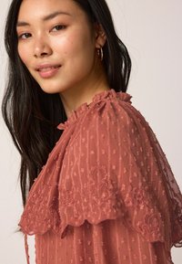 Blouse en dentelle transparente avec un col froncé et des accents en dentelle, présentant une texture en points surélevés et de délicates broderies autour des épaules.