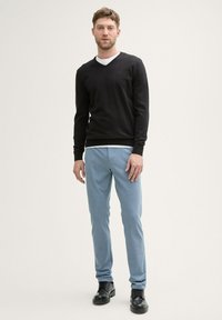 Zwarte v-hals trui over een wit shirt, gecombineerd met lichtblauwe broek en zwarte veterschoenen, staand tegen een neutrale achtergrond.
