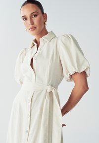 Šviesiai smėlio spalvos marškininė su trumpomis pūstomis rankovėmis, diržu juosmenyje ir lininės tekstūros, su apykakle ir mygtukų juosta priekyje.