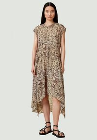 Ženska stoji v brezrokavnih obleki z živalskim potiskom z asimetričnim robom in črnimi ravnimi sandali s trakovi.