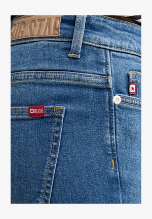 Vaqueros de denim azul con un lavado claro, costuras amarillas y etiquetas rojas en los bolsillos traseros, con un clásico diseño de cinco bolsillos.