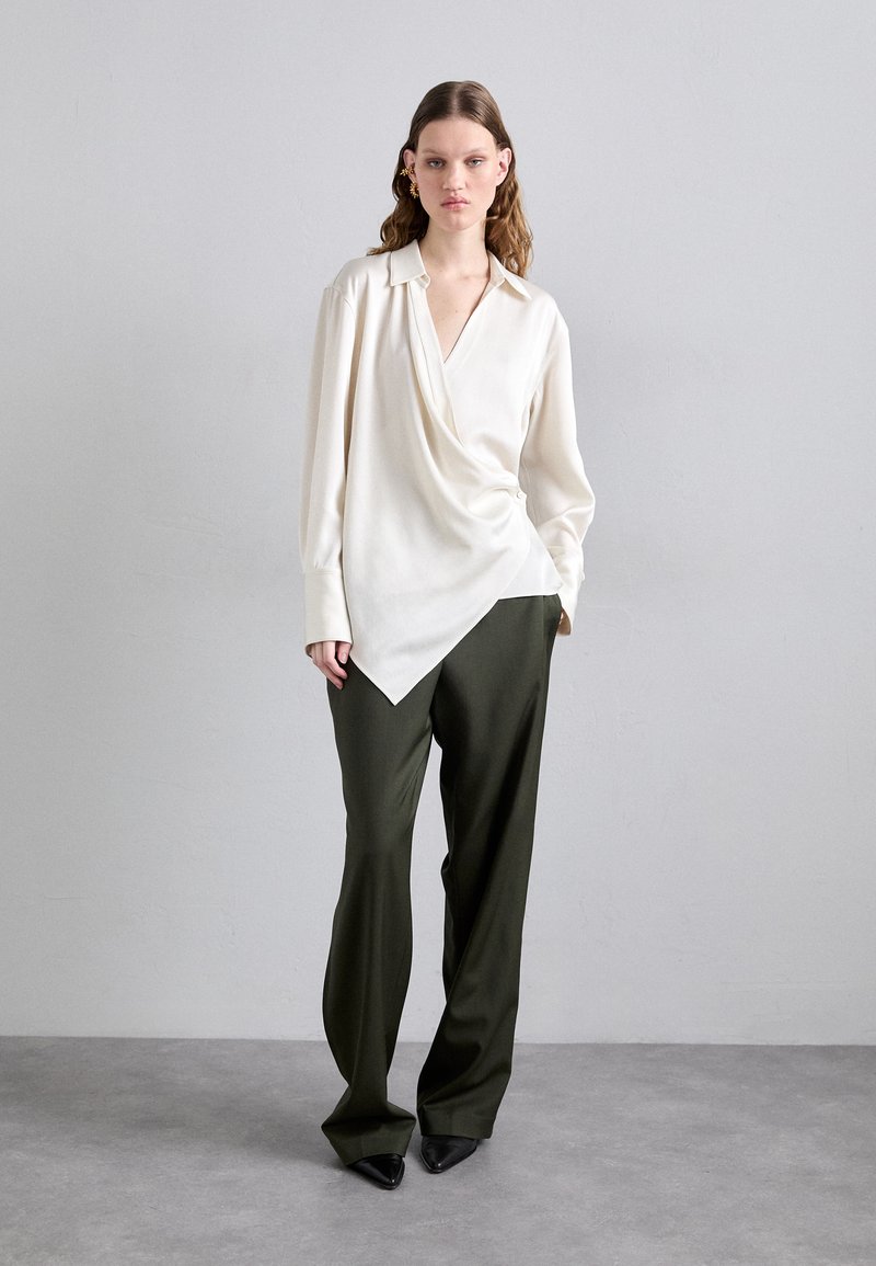 White wrap top with long sleeves and a deep V-neck, paired with olive green wide-leg trousers and pointed black shoes.