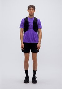 Μαύρο αμάνικο τζάκετ για τρέξιμο πάνω από μωβ μπλουζάκι, συνδυασμένο με μαύρο σορτς και παπούτσια. Το τζάκετ διαθέτει ρυθμιζόμενους ιμάντες και ελαφρύ ύφασμα.