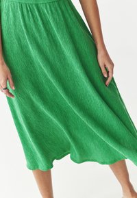 Grünes Midikleid aus strukturiertem Stoff mit fließender Form. Zu den Details gehören ein geraffter Taillenbereich und ein gewellter Saum.