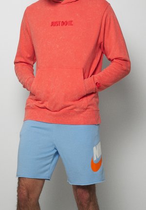 Homme portant un sweat à capuche corail avec le texte « JUST DO IT. » et un short Nike bleu clair arborant un grand logo orange et blanc, mains dans la poche du sweat.