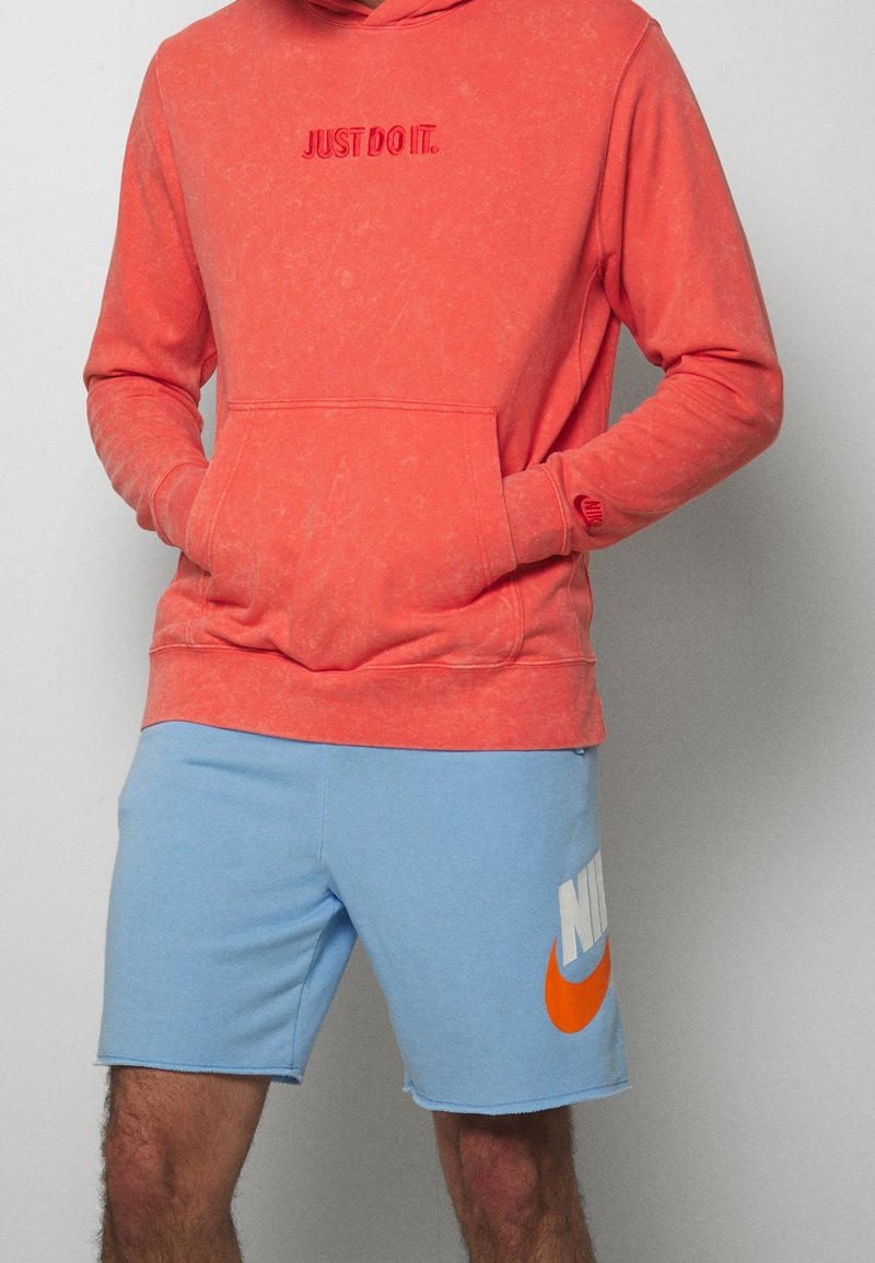 Homme portant un sweat à capuche corail avec le texte « JUST DO IT. » et un short Nike bleu clair arborant un grand logo orange et blanc, mains dans la poche du sweat.