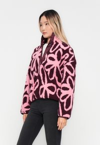 Pink and burgundy fleece pullover with floral pattern, high collar, and snap buttons, paired with black fitted jeans. Soft, textured fabric.