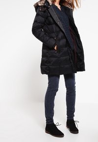 Svart vadderad kappa med fuskpäls-hette, dragkedja och knappstängning, två sidofickor; kombinerad med blå byxor och svarta ankelstövlar.