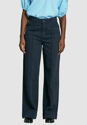 Person trägt hochtaillierte, weit geschnittene dunkle Hose mit vorderen Nahtdetails, kombiniert mit einem blau-weiß gestreiften Hemd und schwarzen Schuhen.